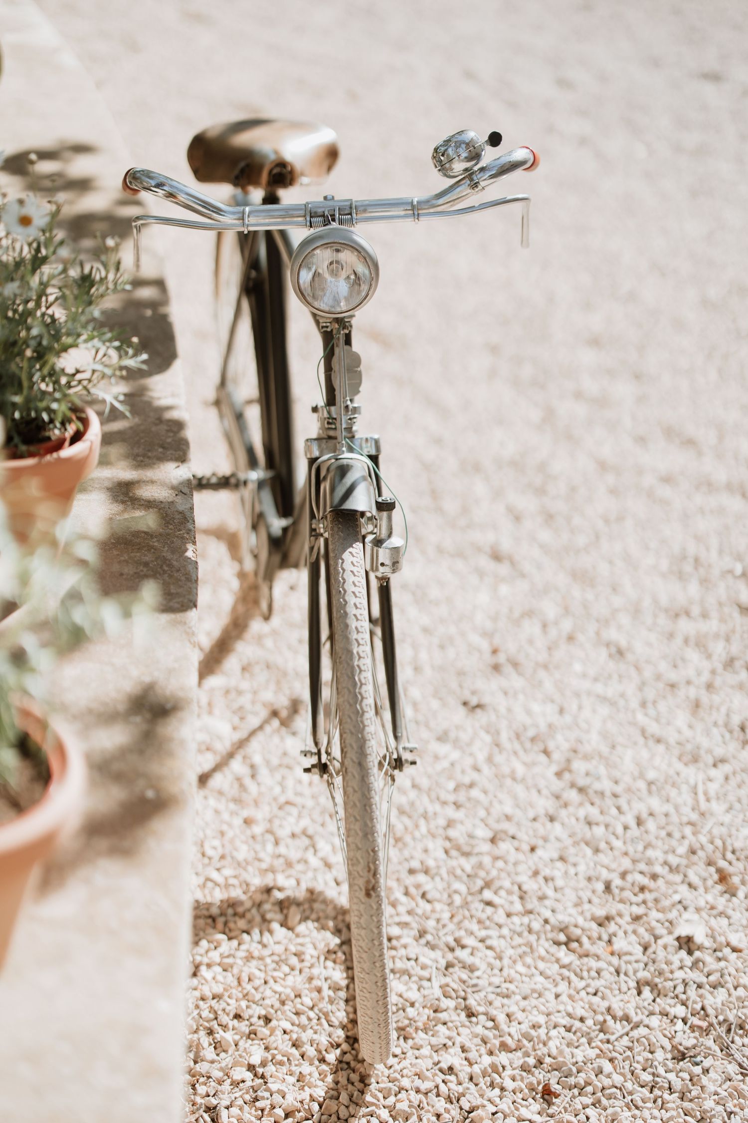 Close-up view of a vintage bicycle handlebars and front wheel on gravel ground with potted plants in soft muted tones.