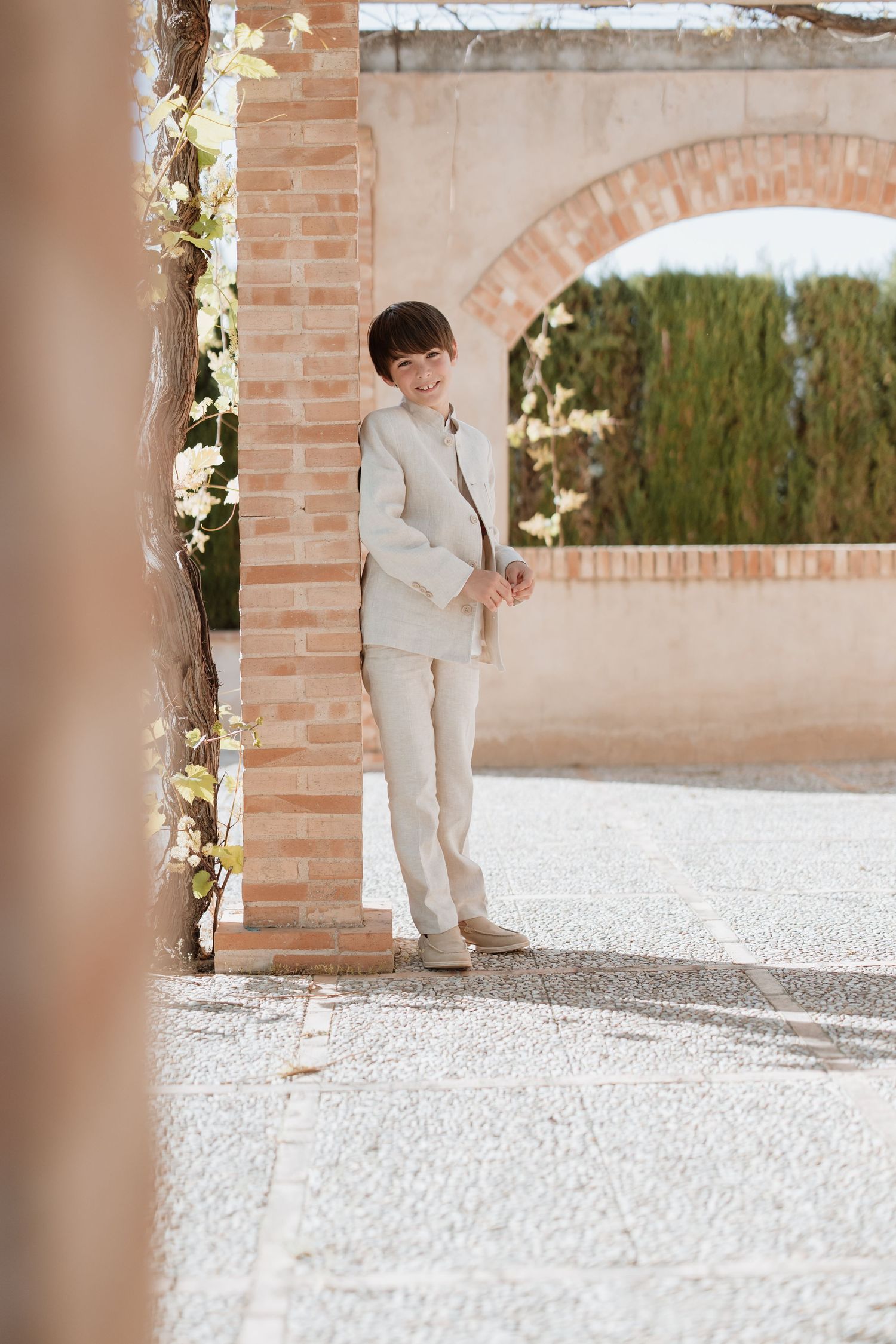 Someone wearing a light beige suit stands in an elegant archway decorated with white flowers and climbing vines.