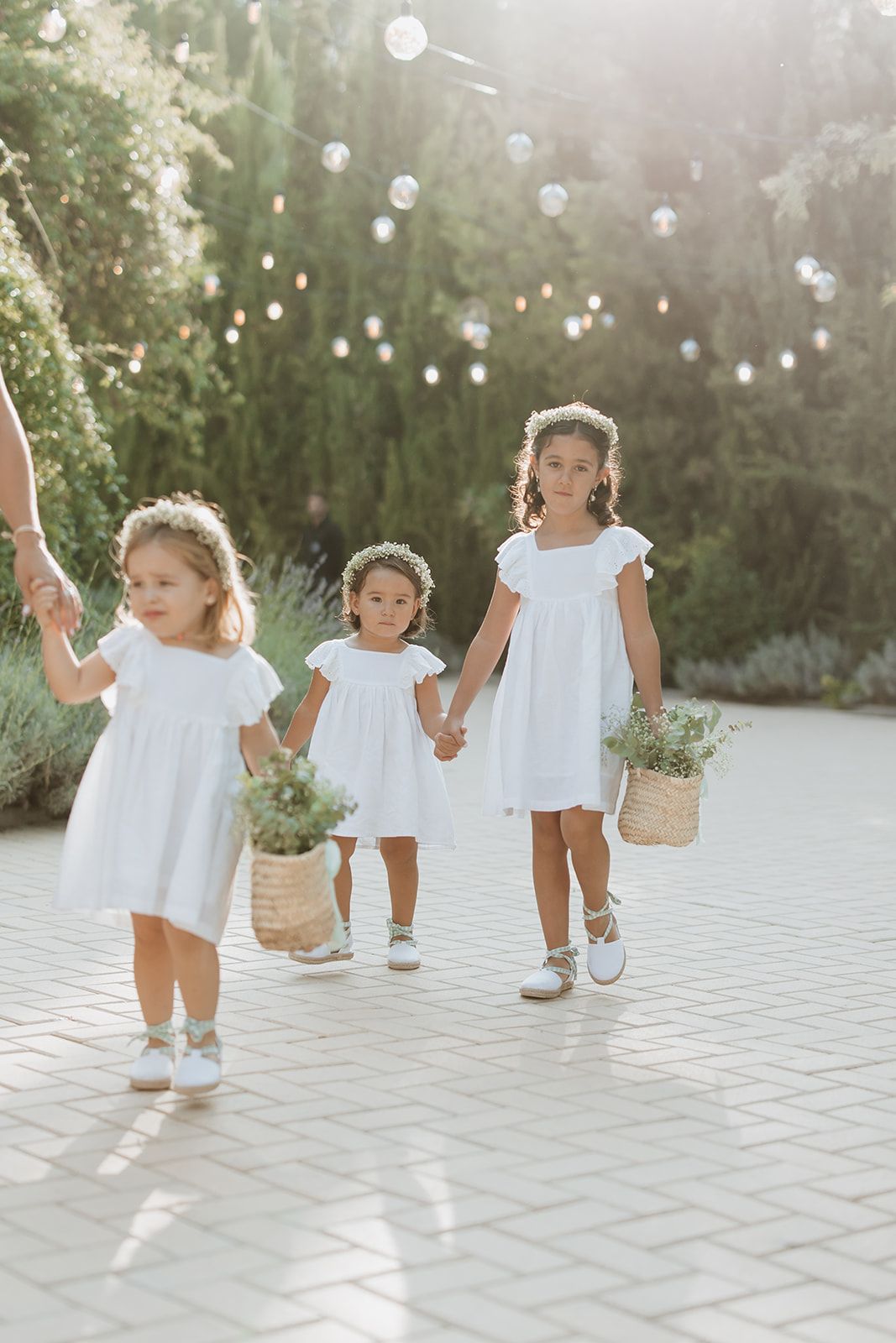 Young flower girls in white dresses carry baskets along a stone pathway under twinkling lights.