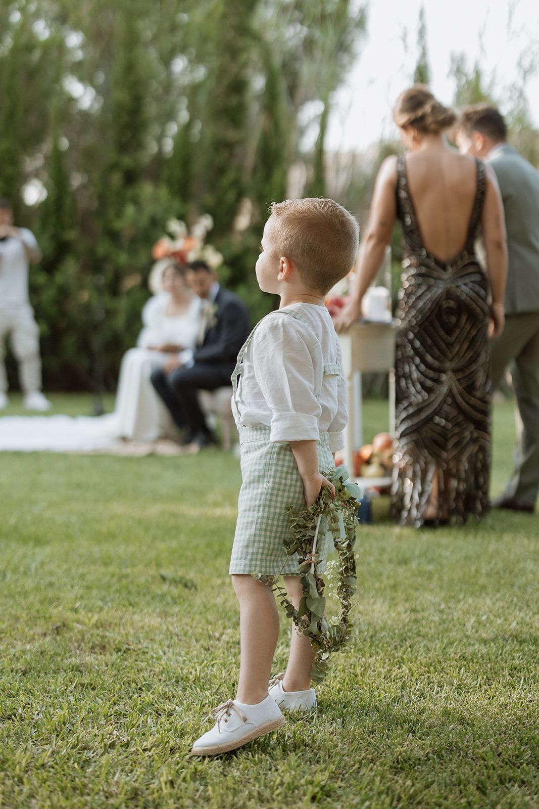 Young ring bearer in checkered shorts and white shirt standing on grass during outdoor wedding.