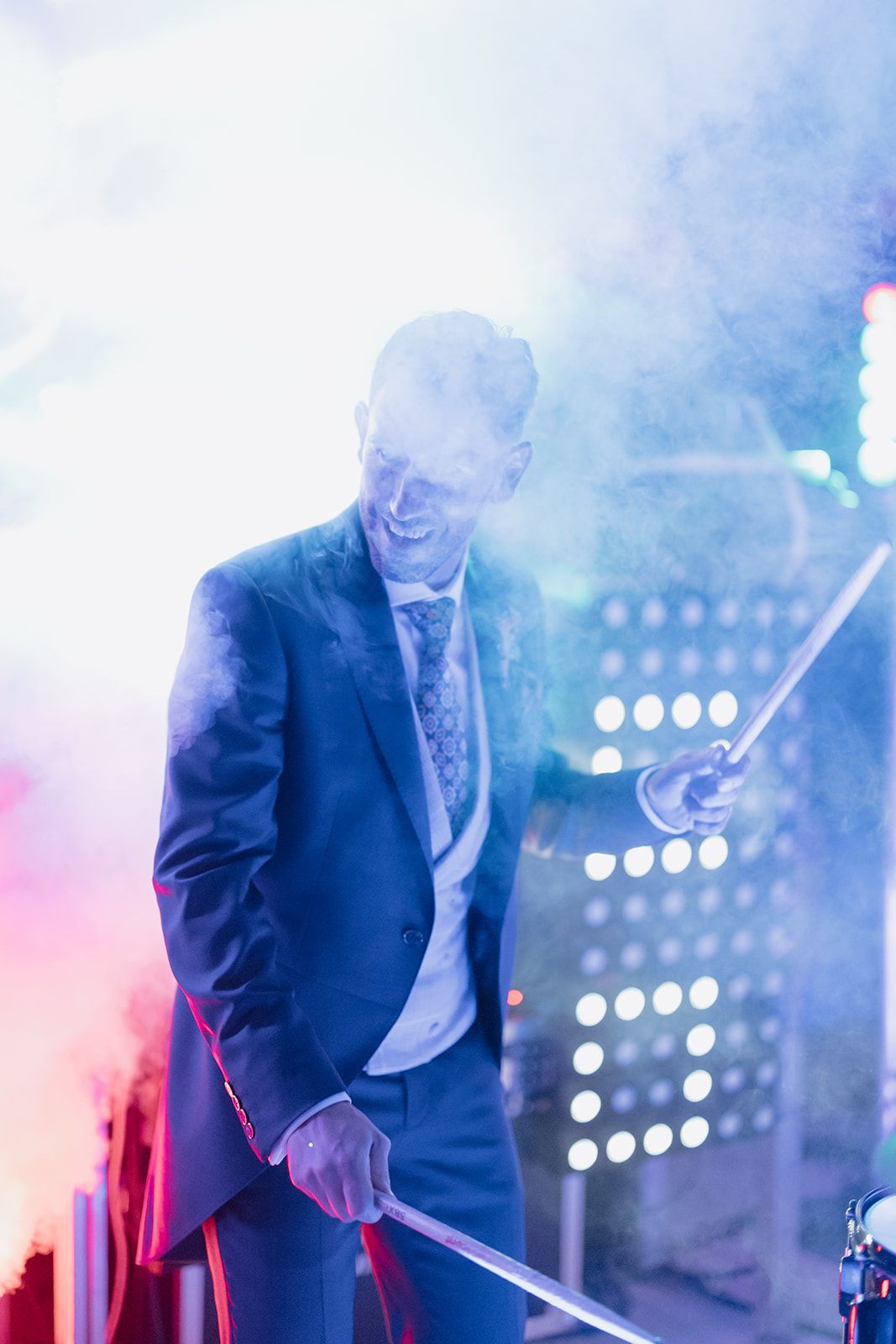 Suited performer on stage surrounded by colorful smoke and stage lighting.