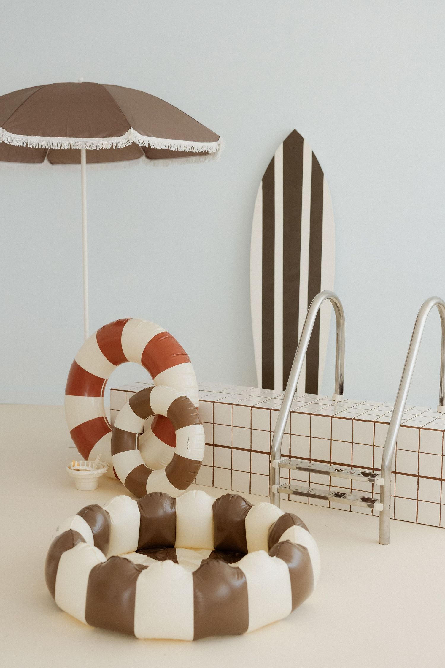 Brown and cream striped inflatable pool rings near a mini pool with metal rails and a striped surfboard under a fringed umbrella.