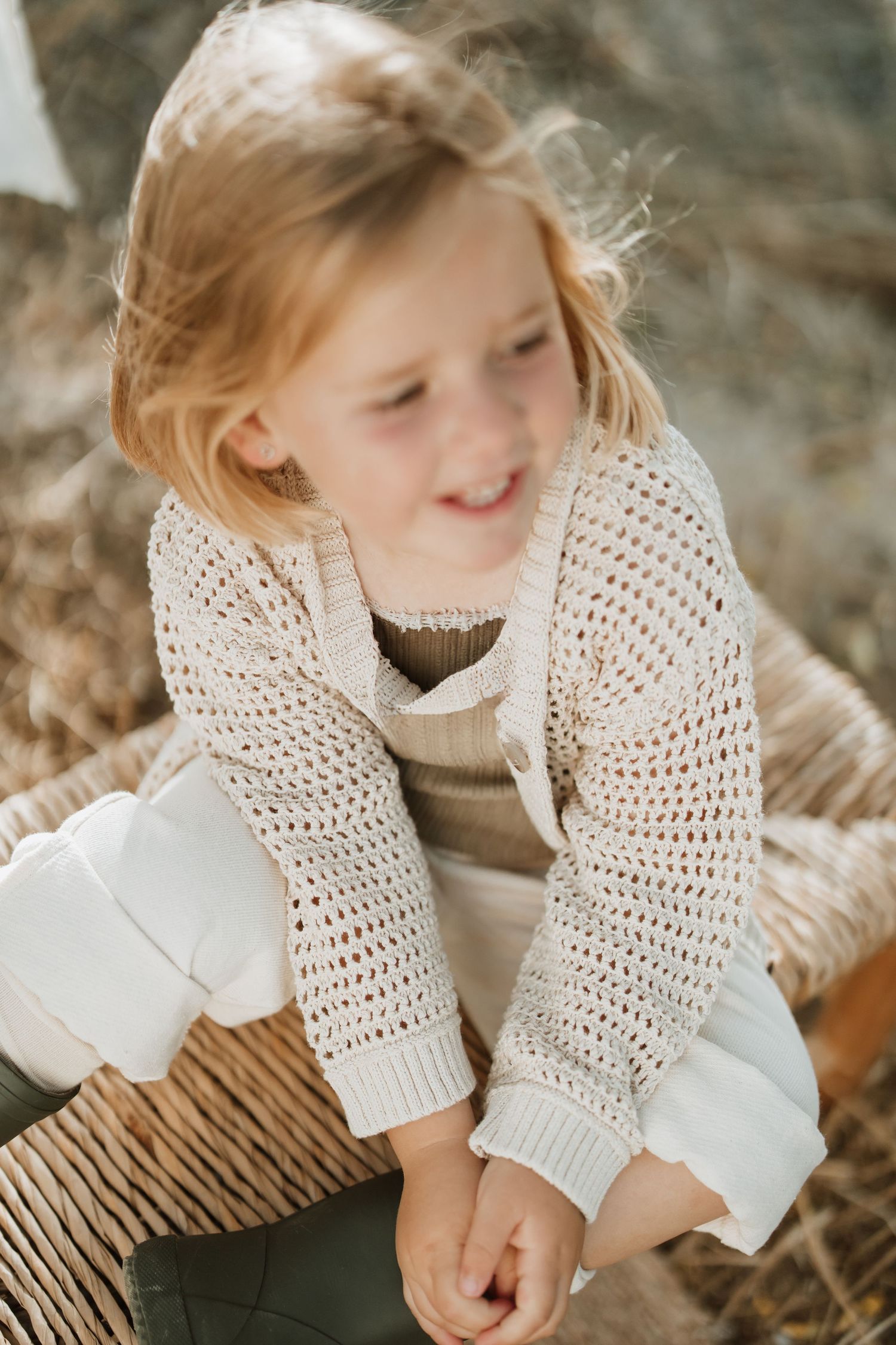 Child in knit cardigan sitting on woven bench outdoors during golden hour photoshoot.