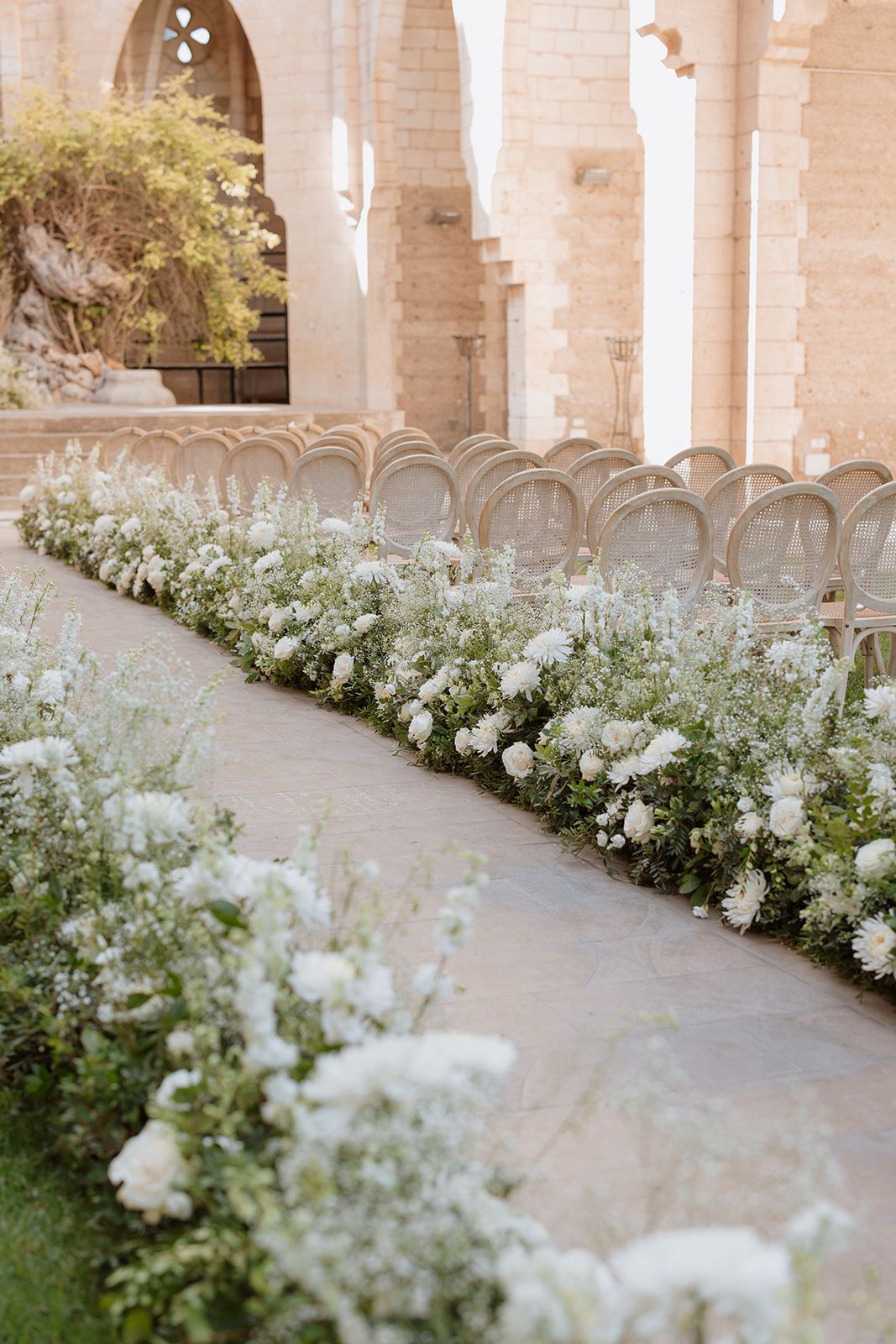 Elegant curved chairs line a stone path bordered by white floral arrangements and gothic arches.