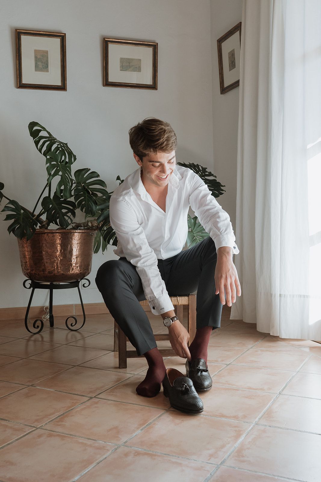 Person in white shirt and dark pants sitting on chair next to potted monstera plant.