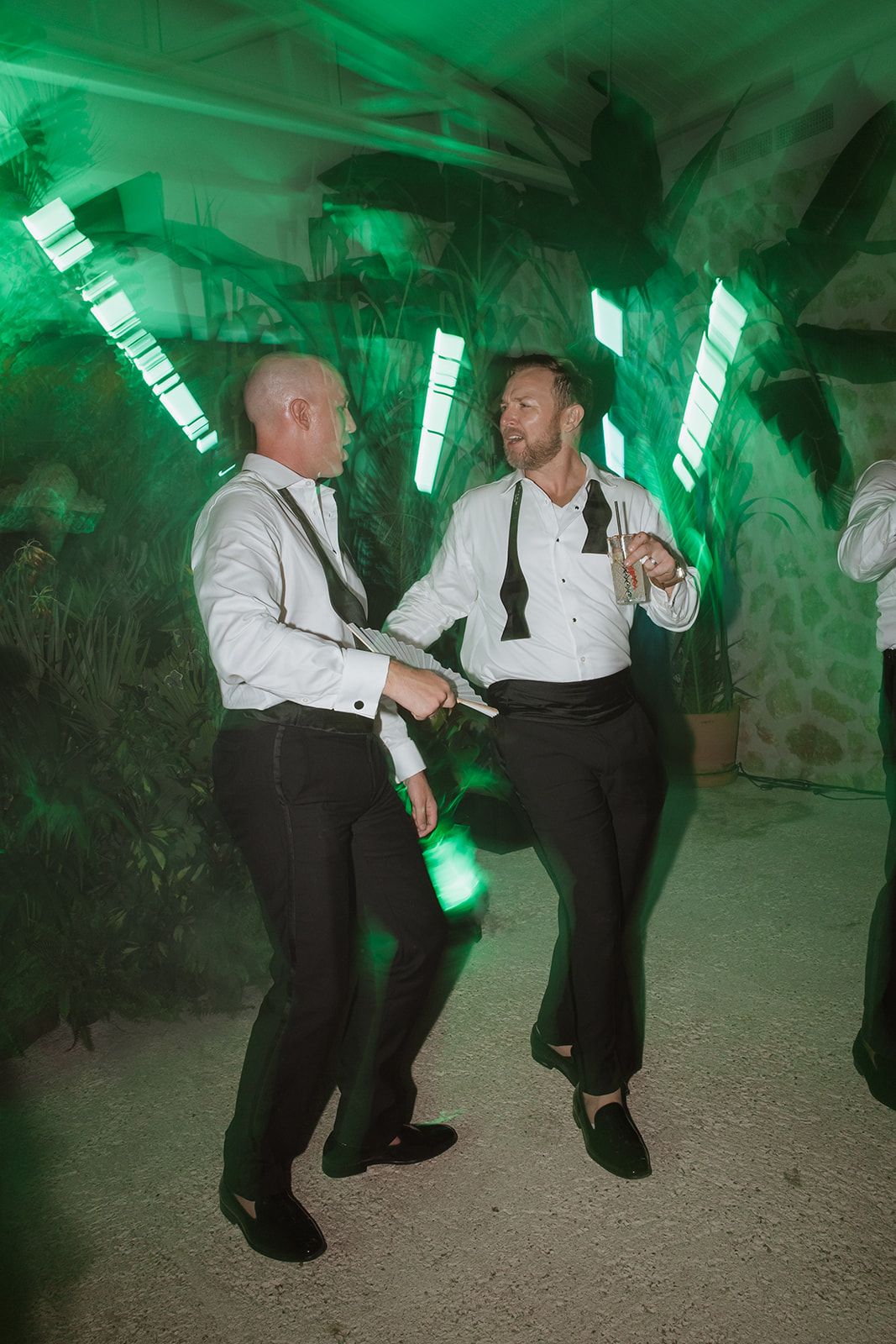Two men dancing energetically under green lighting at wedding reception.