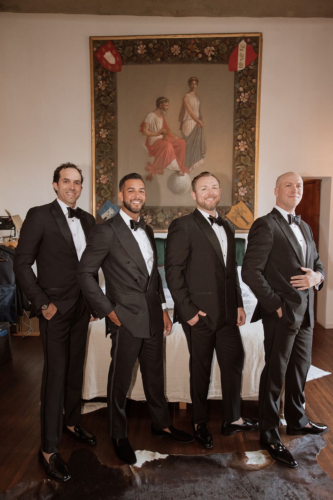 Four groomsmen in black tuxedos pose together in front of a decorative framed artwork.