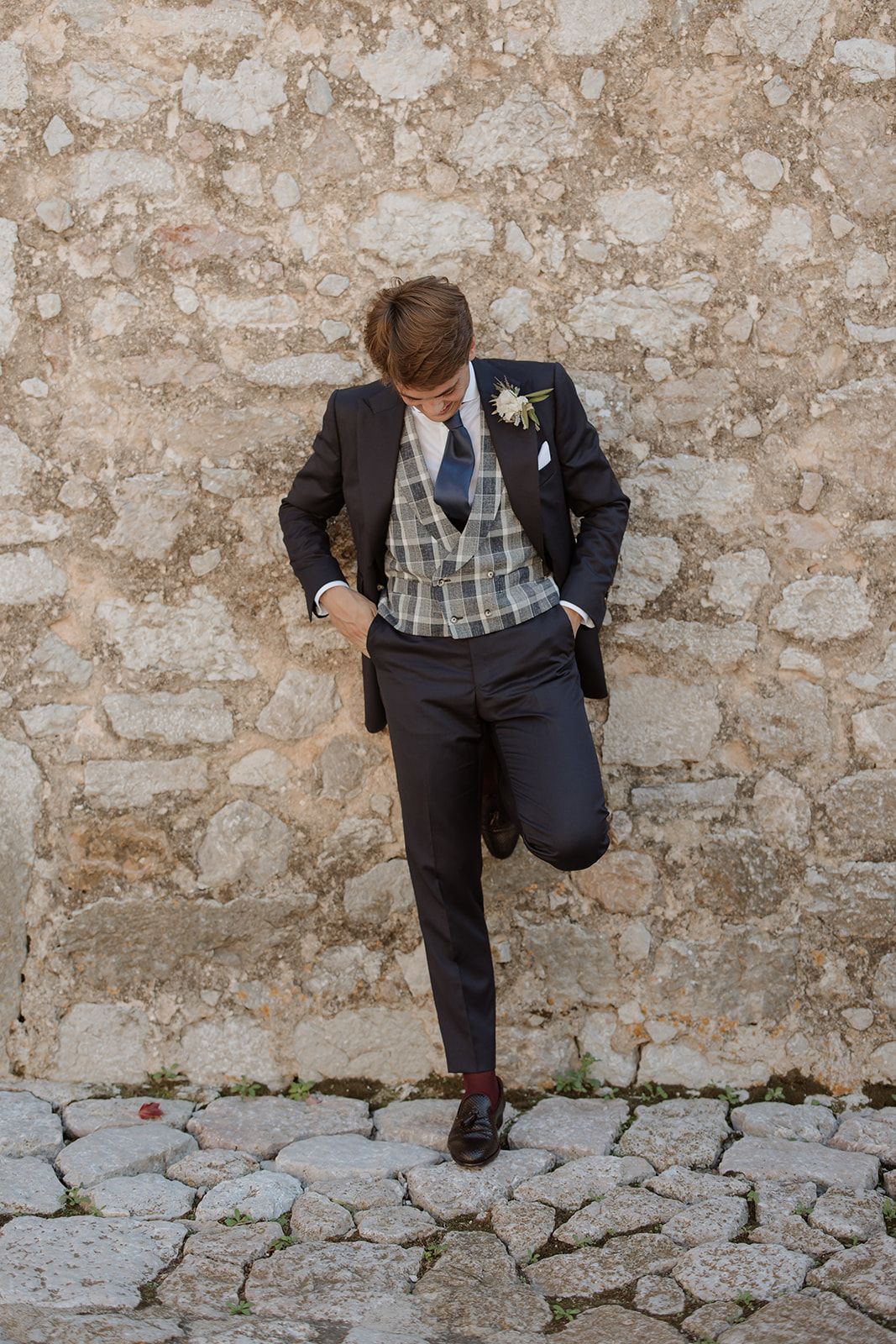 Groom in dark suit with plaid vest poses against rustic stone wall during wedding photoshoot.