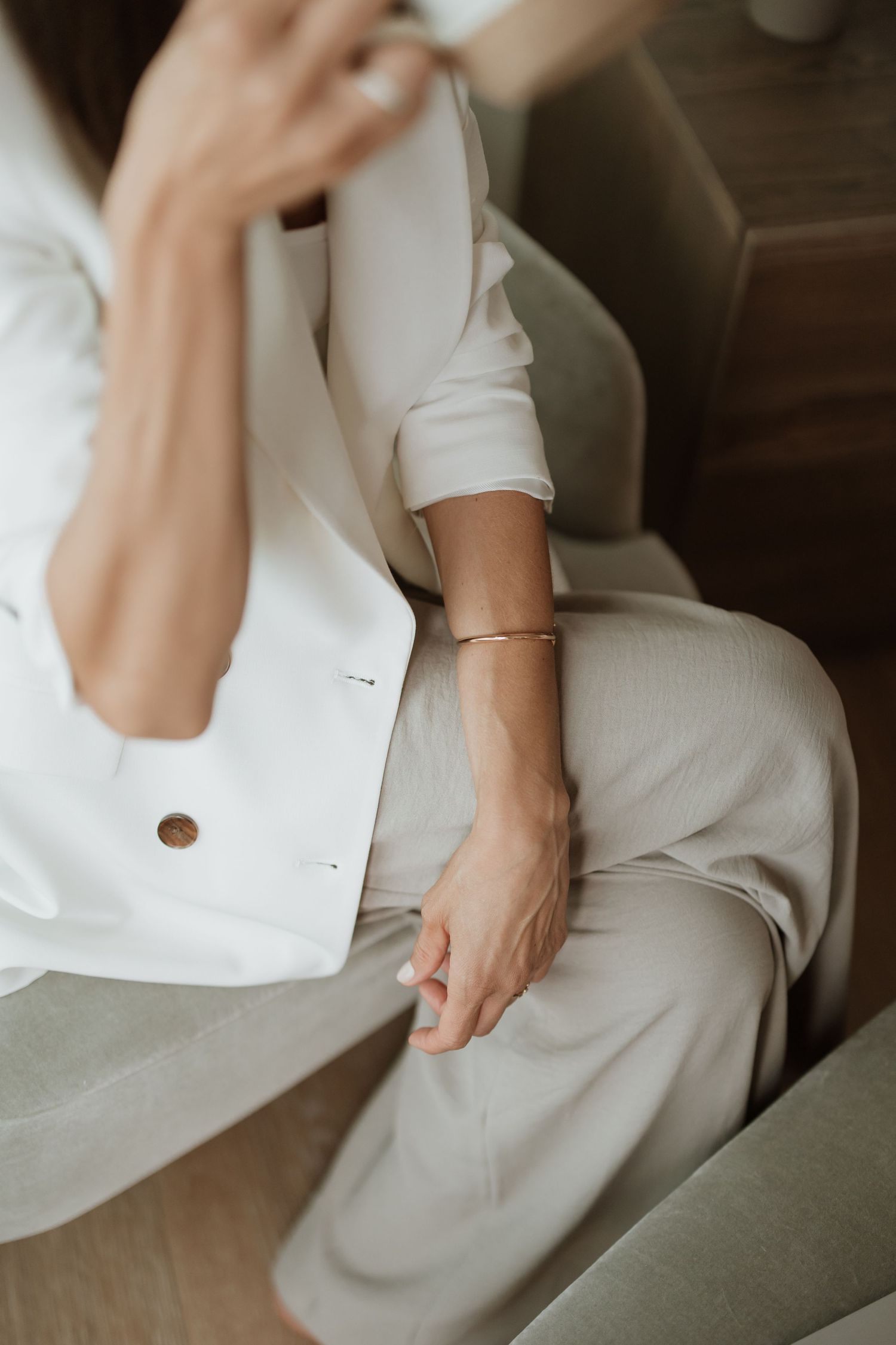 Detail shot of white blazer sleeve and delicate gold bracelet on grey chair.