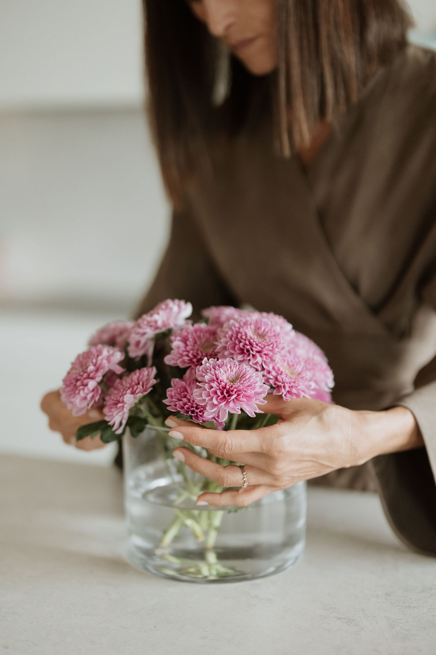 Hands gently arranging pink chrysanthemums in clear glass vessel.