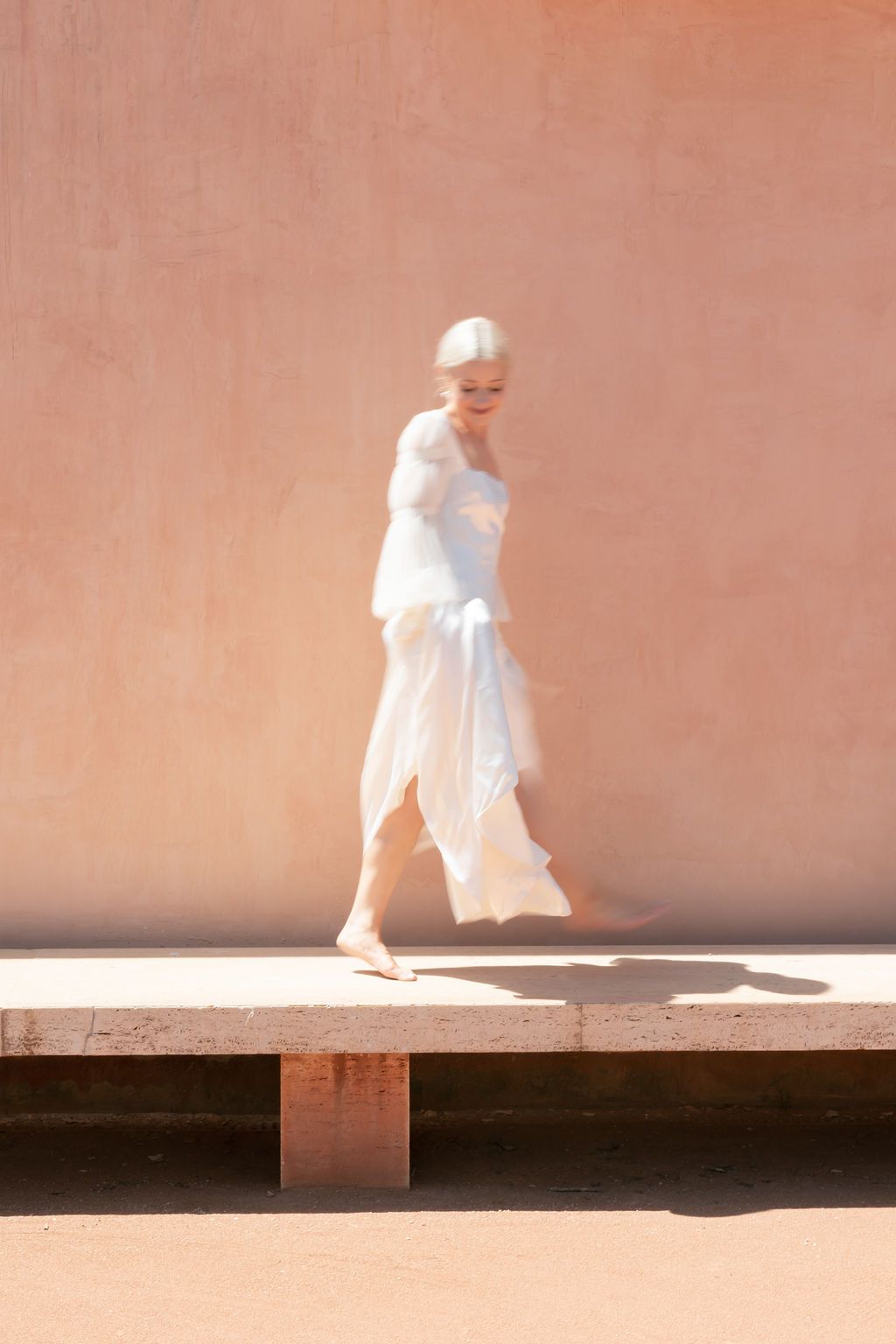 Ethereal portrait of a person in flowing white dress against a pink wall.