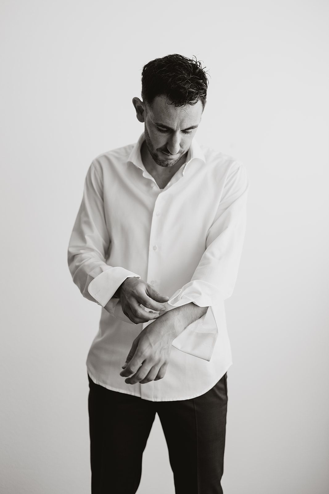 A person in formal attire smiles while fastening their white dress shirt.