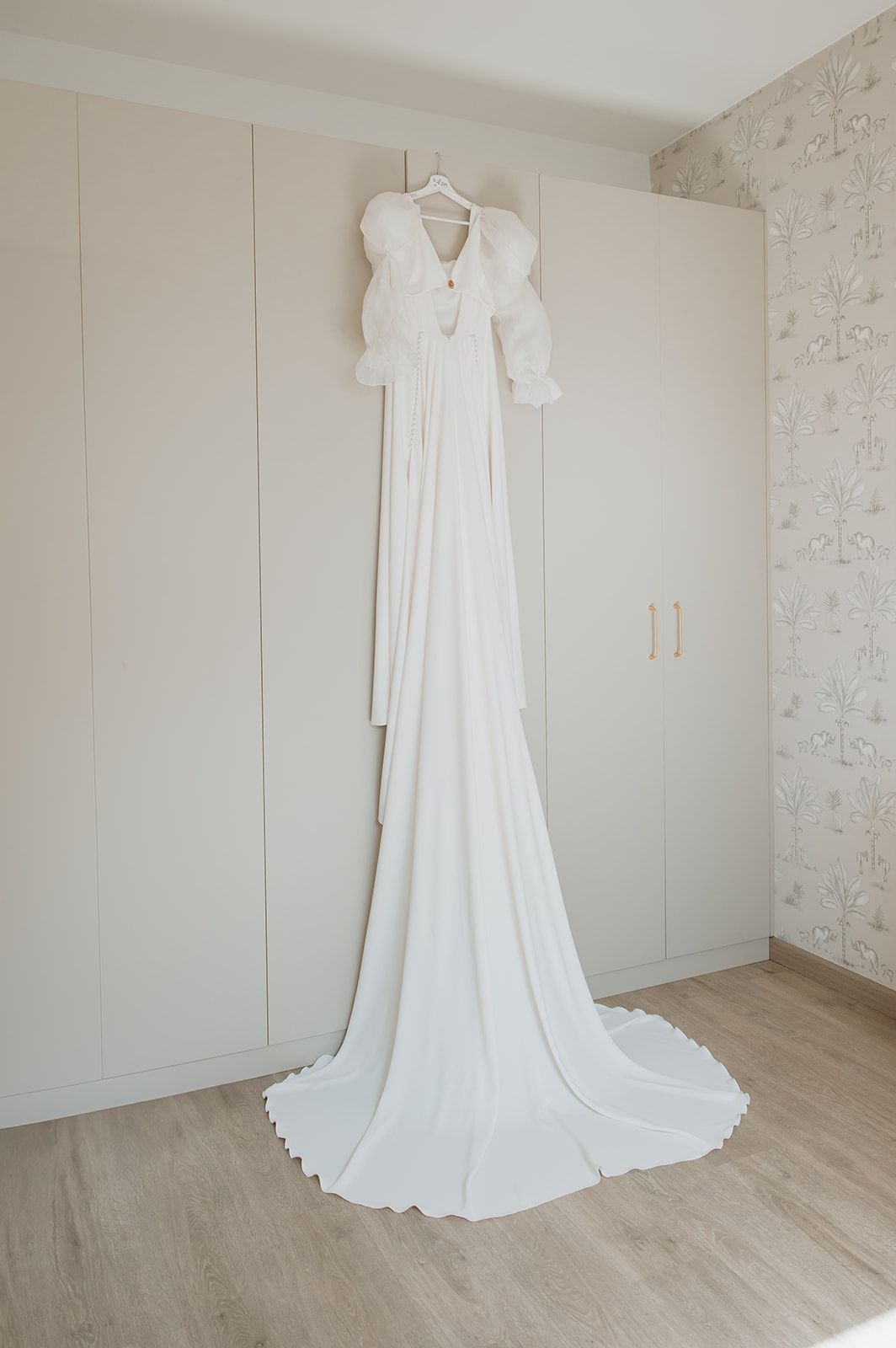 Elegant white wedding gown with long sleeves and flowing train hanging against a light wall.