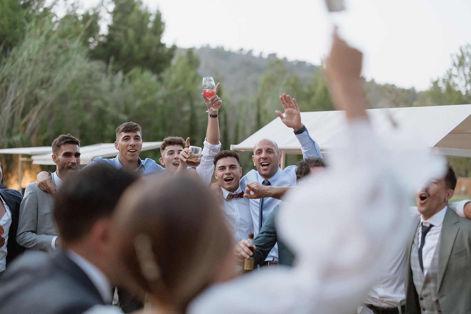 Wedding guests raise their glasses in a celebratory toast outdoors.