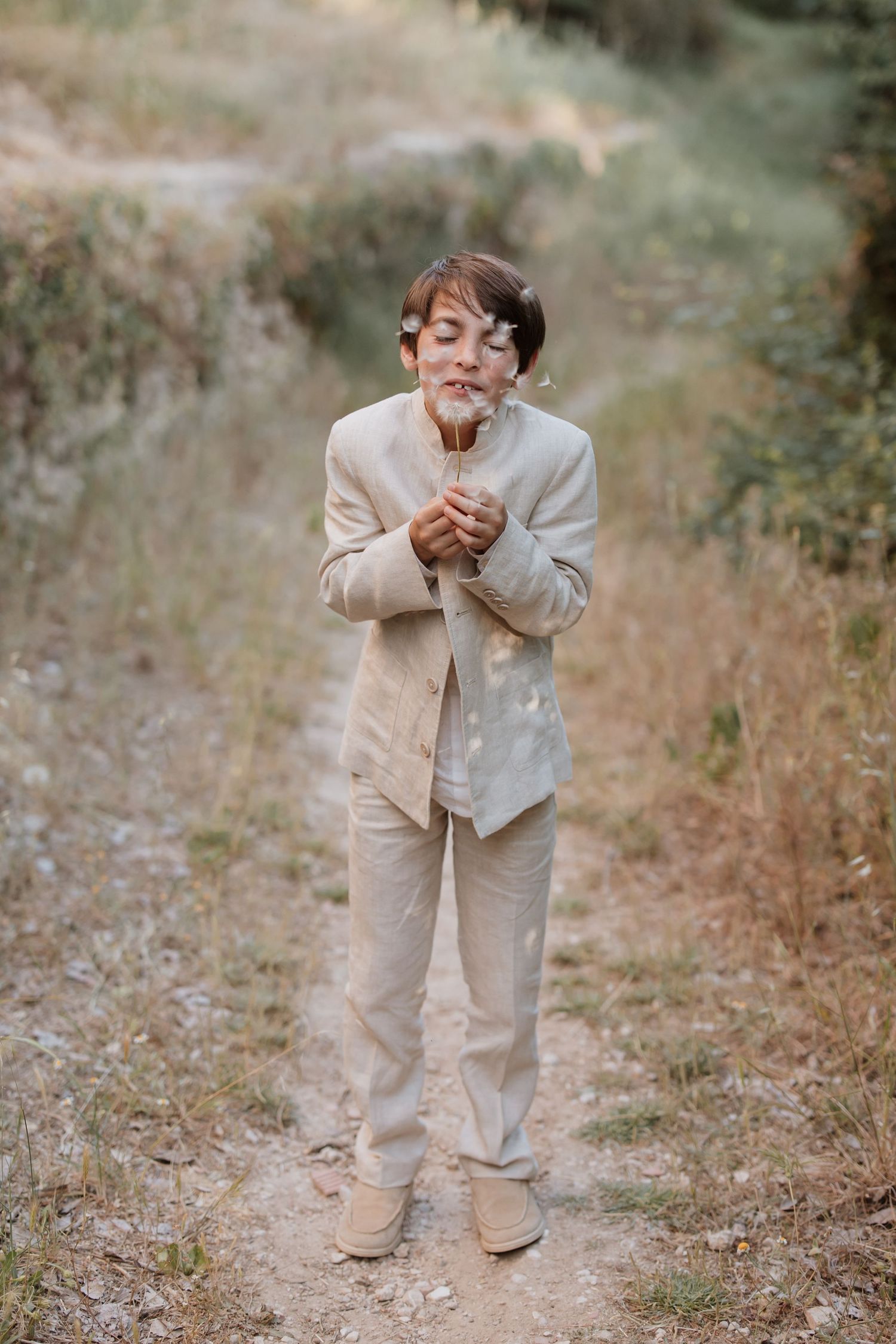 A person wearing a beige linen suit and boots stands on a dirt path while joyfully blowing on a dandelion.