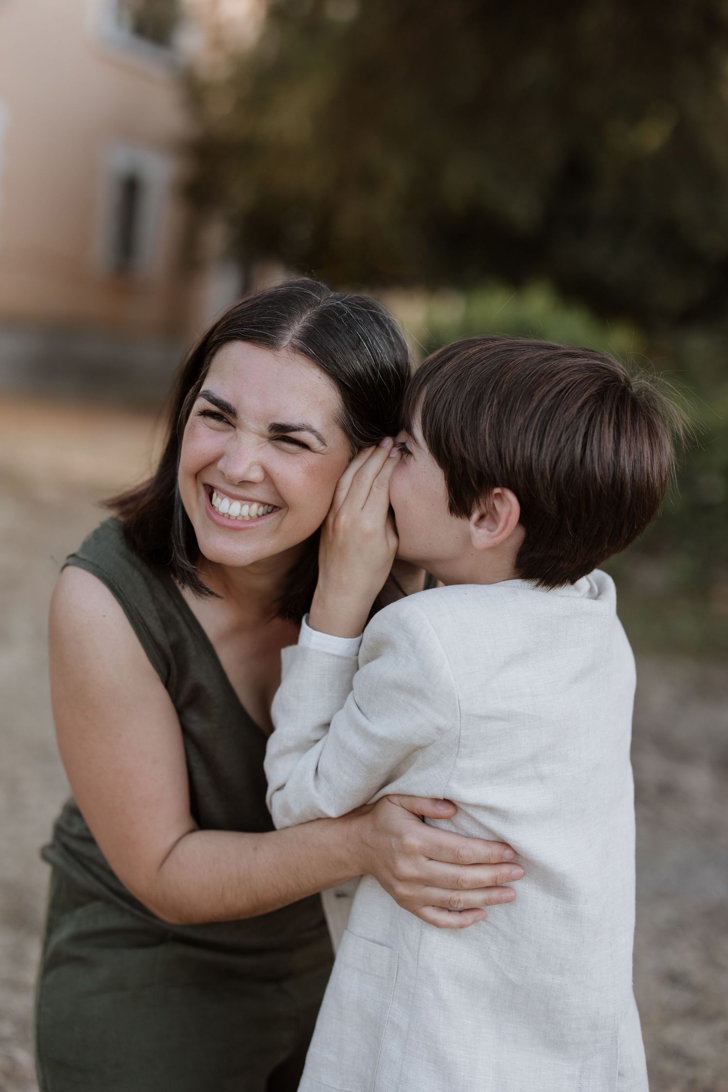 A heartwarming candid moment shared between a young child whispering and a smiling woman in a green dress outdoors.