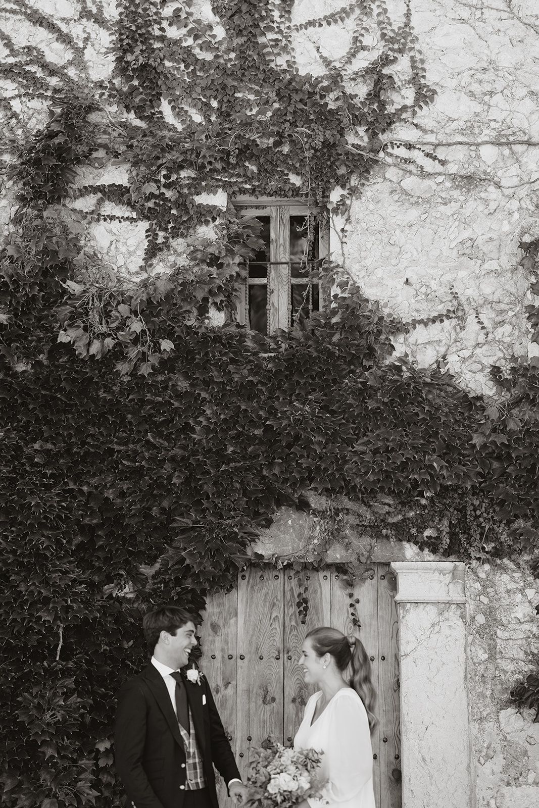 A couple shares an intimate moment in front of a vine-covered wooden door.