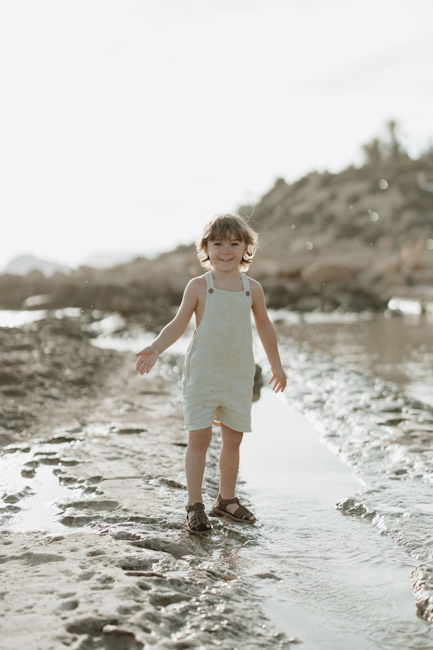 A little one in a light-colored romper explores the water's edge on a rocky beach.