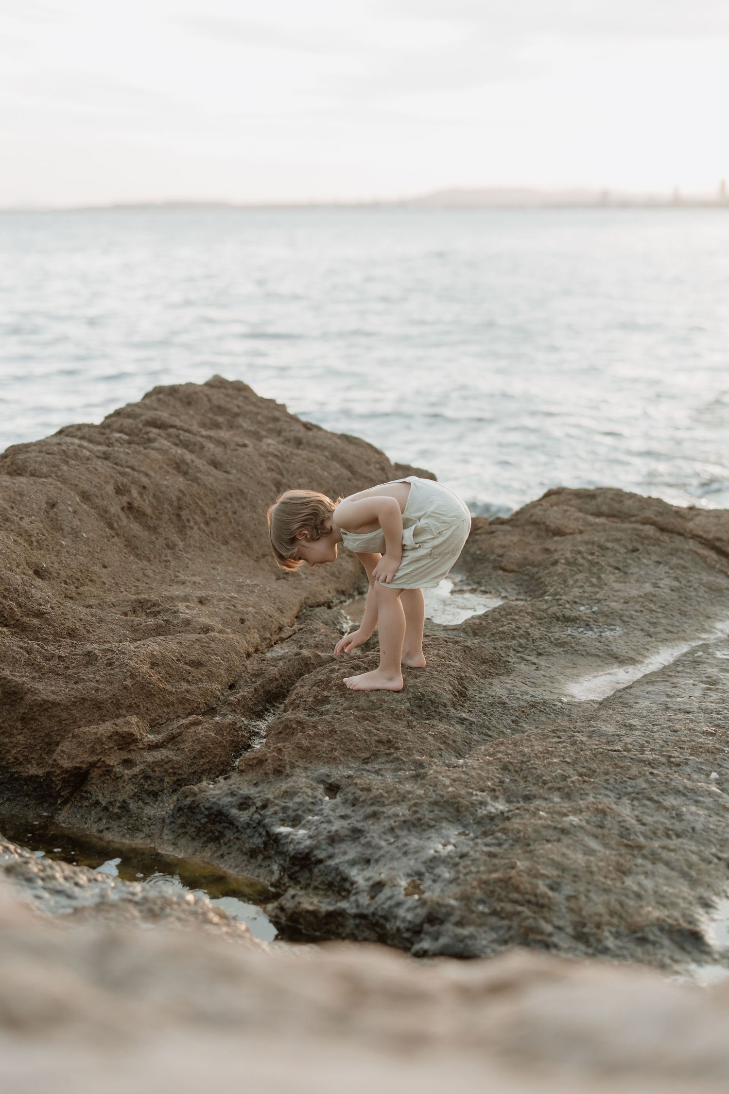 Small child exploring rocky beach formation near ocean at sunset.