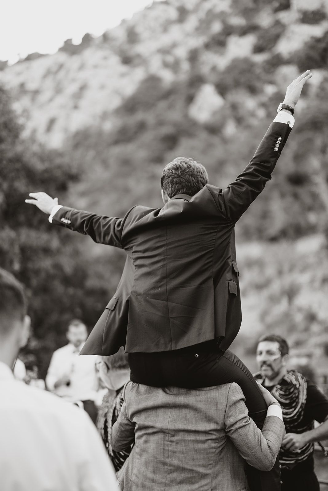 A wedding guest rides on shoulders with arms outstretched during outdoor festivities.