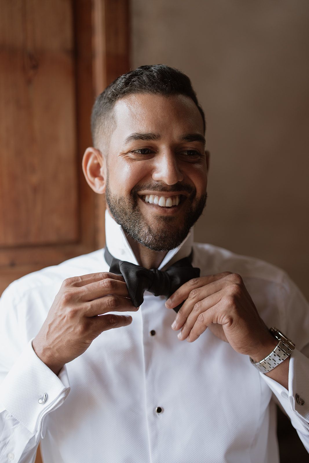 A groom in a crisp white tuxedo shirt adjusts his black bow tie with a bright smile.