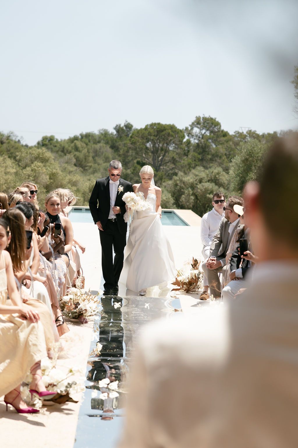 Wedding ceremony procession on mirrored aisle with guests seated in neutral colors.