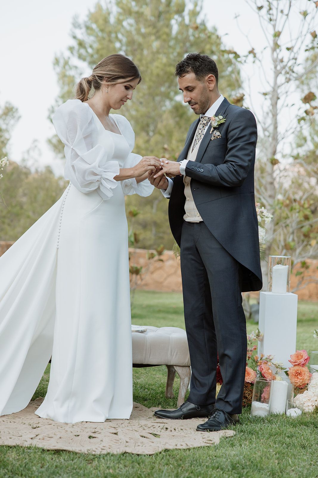 A wedding couple exchanging rings during their outdoor ceremony surrounded by floral decorations.