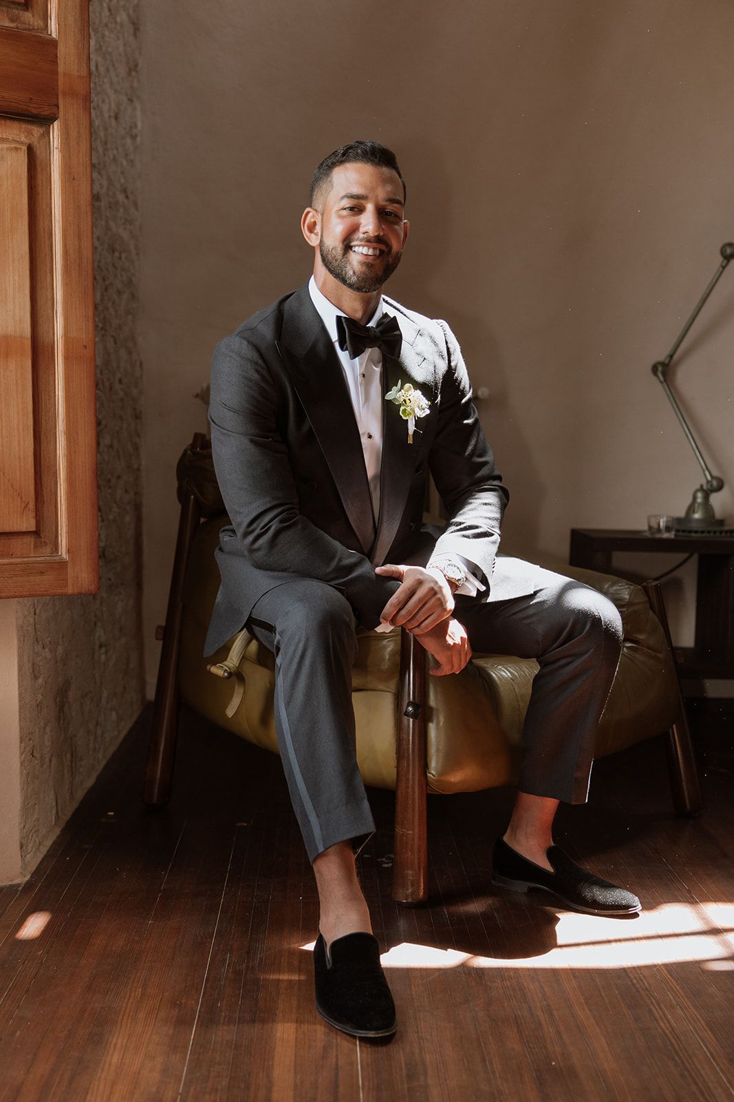 Person in black tuxedo sits confidently on chair in sunlit room.