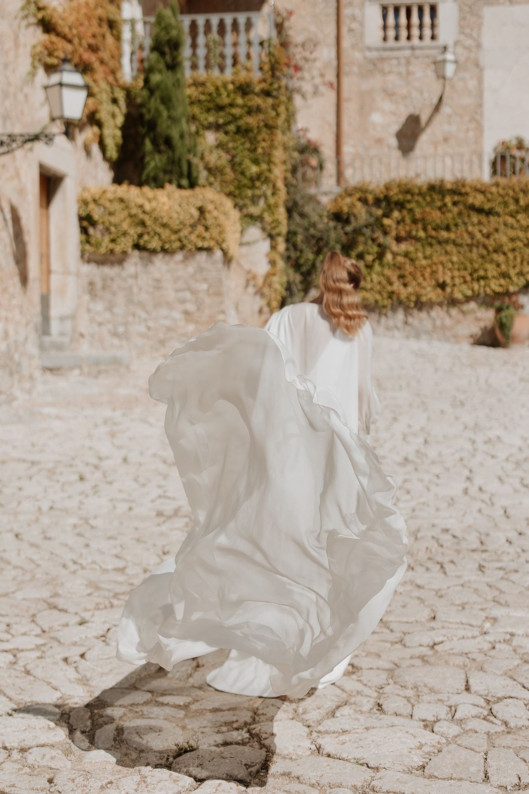 Flowing white wedding dress catches wind in Mediterranean courtyard setting.