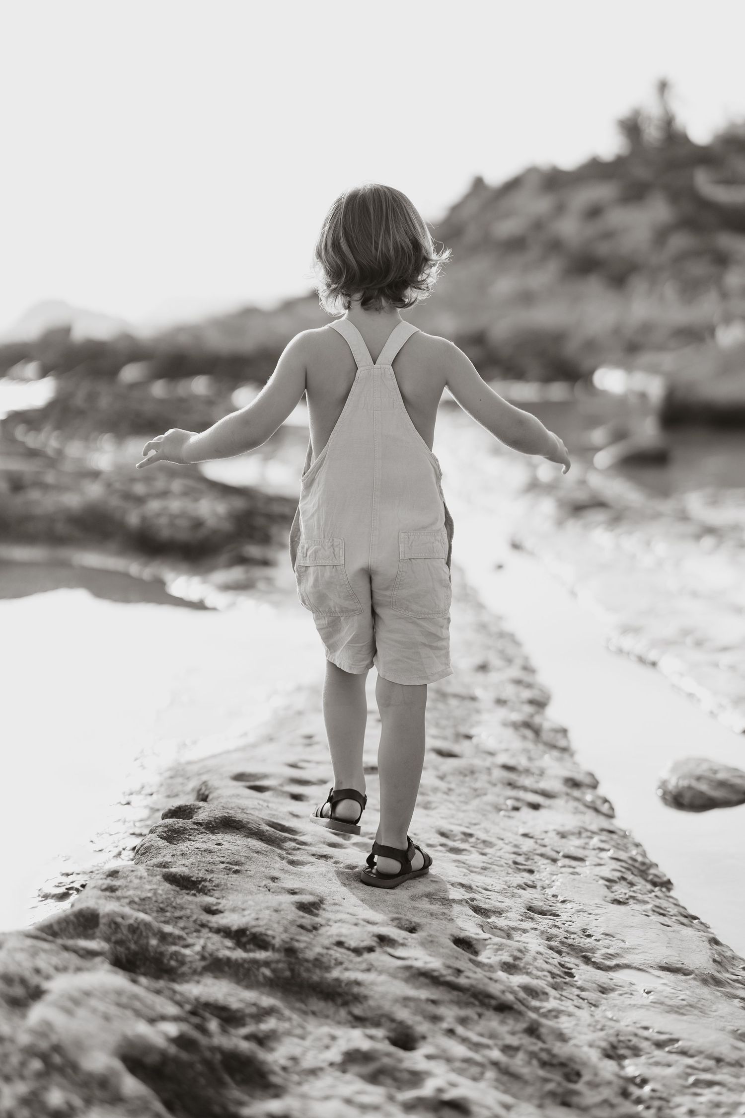 Child in white overalls walking on rocky beach path in black and white.