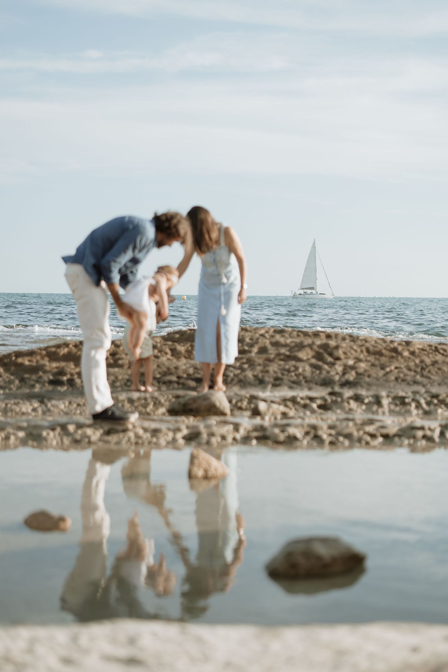 Family looking at reflections in tidepool on rocky coastline with sailboat in background.
