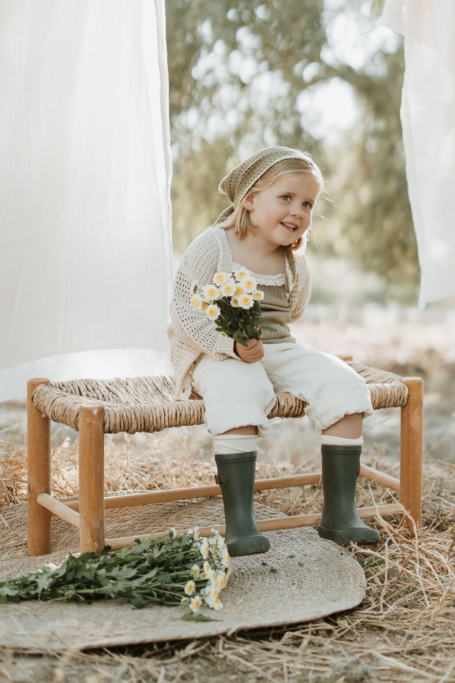 Child in cream sweater and green boots holding flowers while sitting on rustic bench between white curtains.