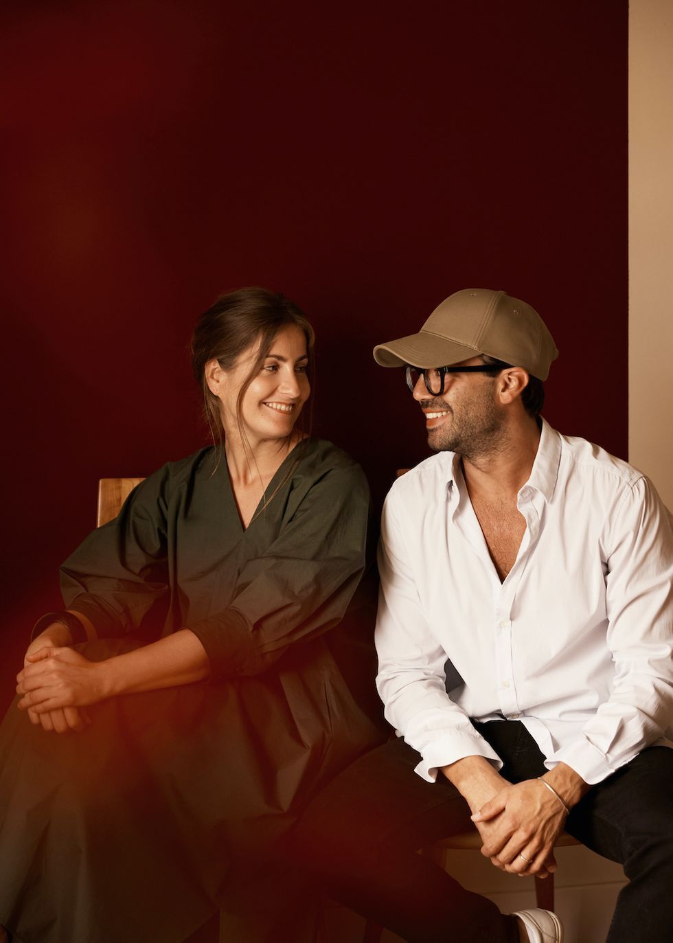 A man in a white shirt and cap smiles at a woman in an olive dress against a deep red background.