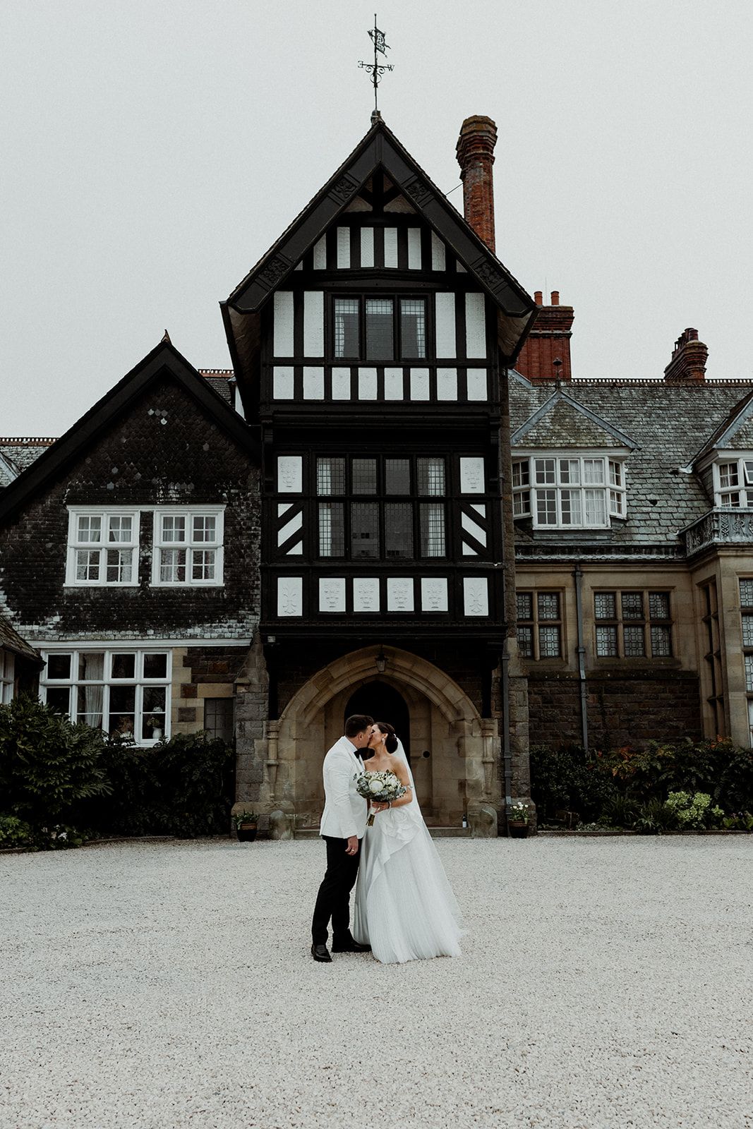 Newlywed couple embracing in front of historic Tudor-style mansion with black and white timber framing and stone archway.