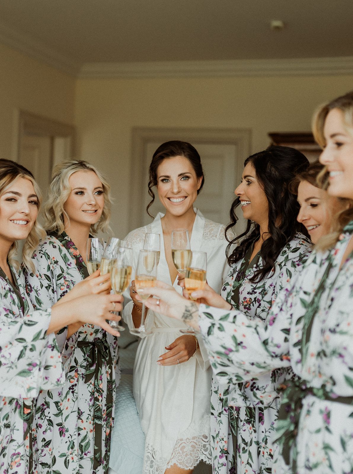 A bridal party in matching floral robes toasts with champagne glasses during wedding preparations.