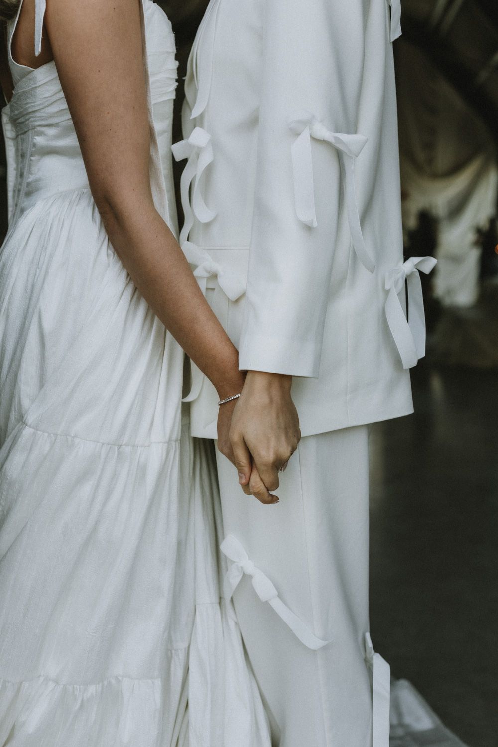 Close-up of two people holding hands while wearing elegant white wedding attire.