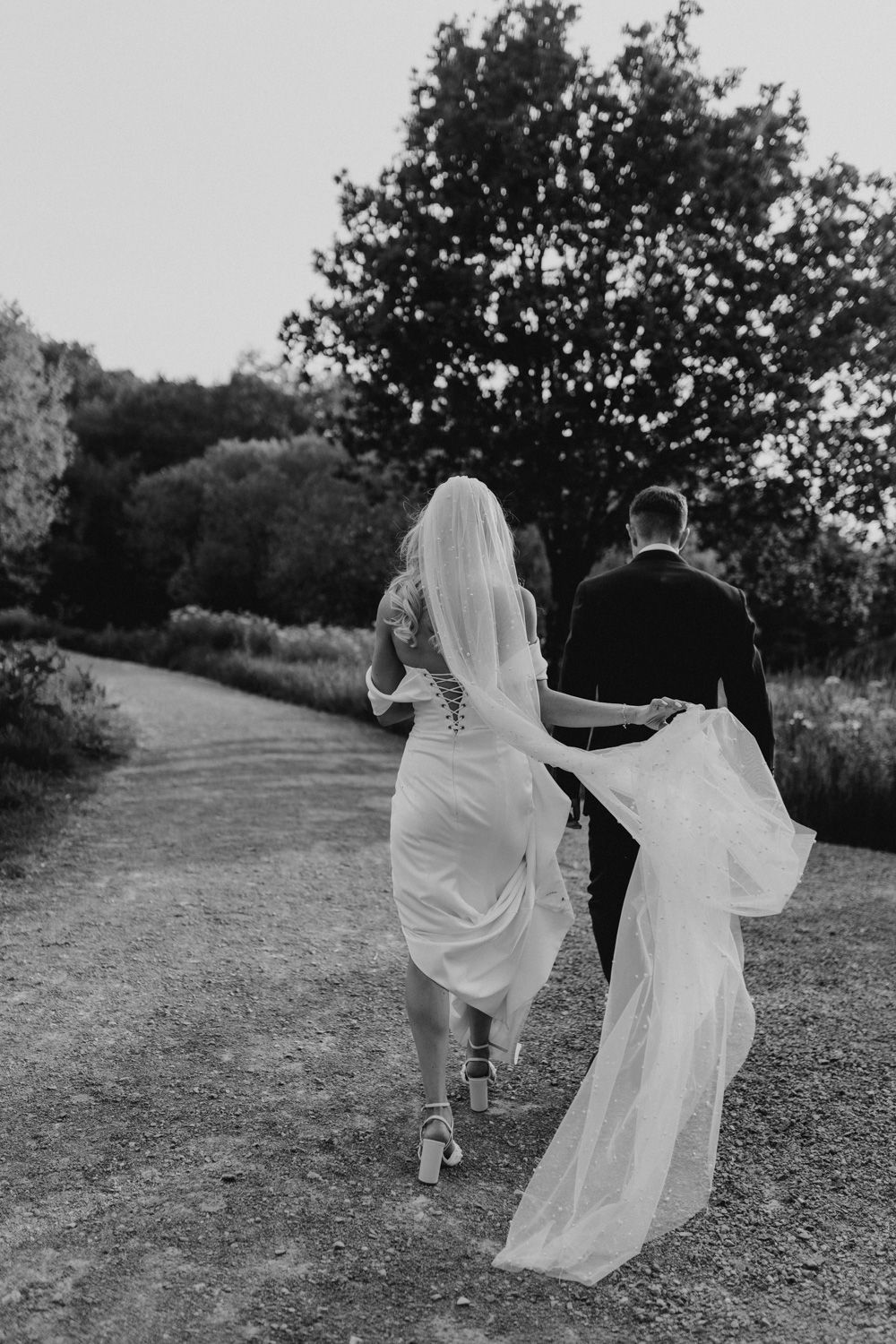 Black and white photo of a couple running playfully on a dirt path with flowing wedding dress.