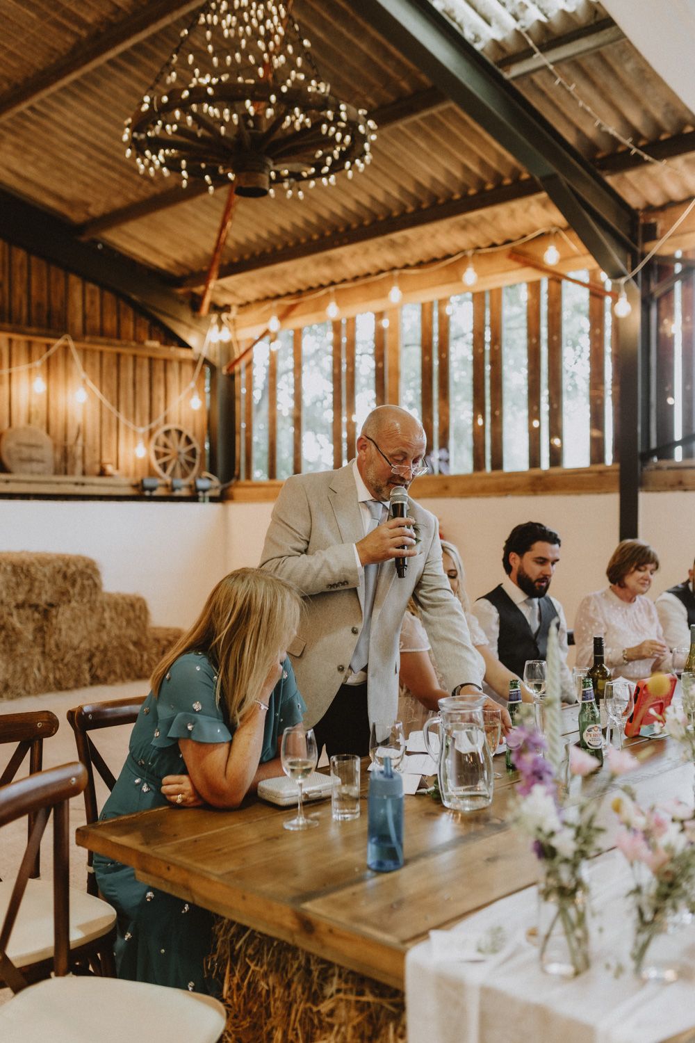 Wedding reception speech in a rustic barn venue with string lights and hay bale seating.
