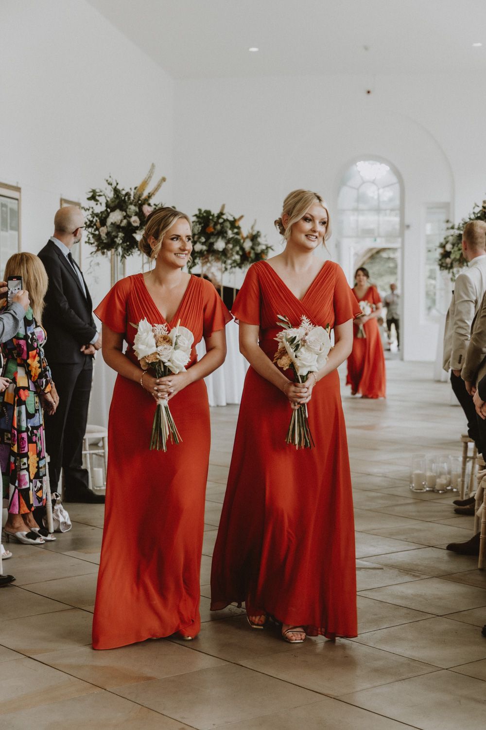 Bridesmaids in floor-length red gowns stand together holding bouquets in a bright indoor venue.