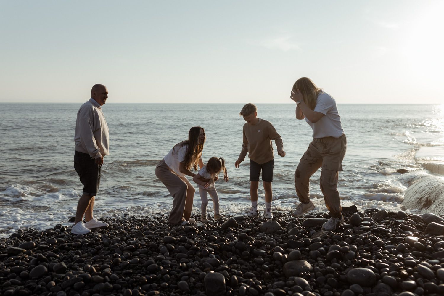 Family plays together on rocky beach near ocean waves at sunset.