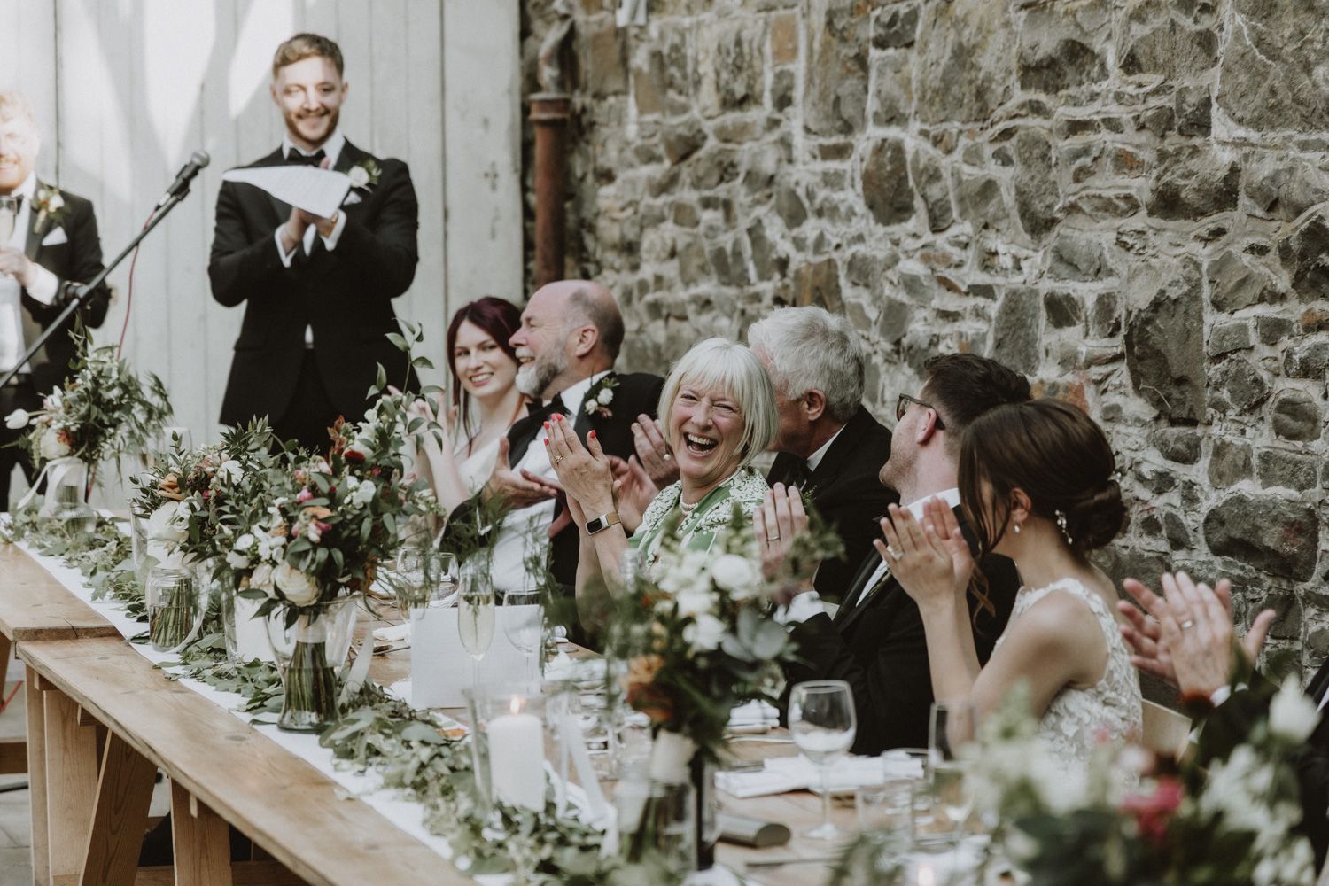 Wedding reception toast with guests applauding at long table decorated with flowers.