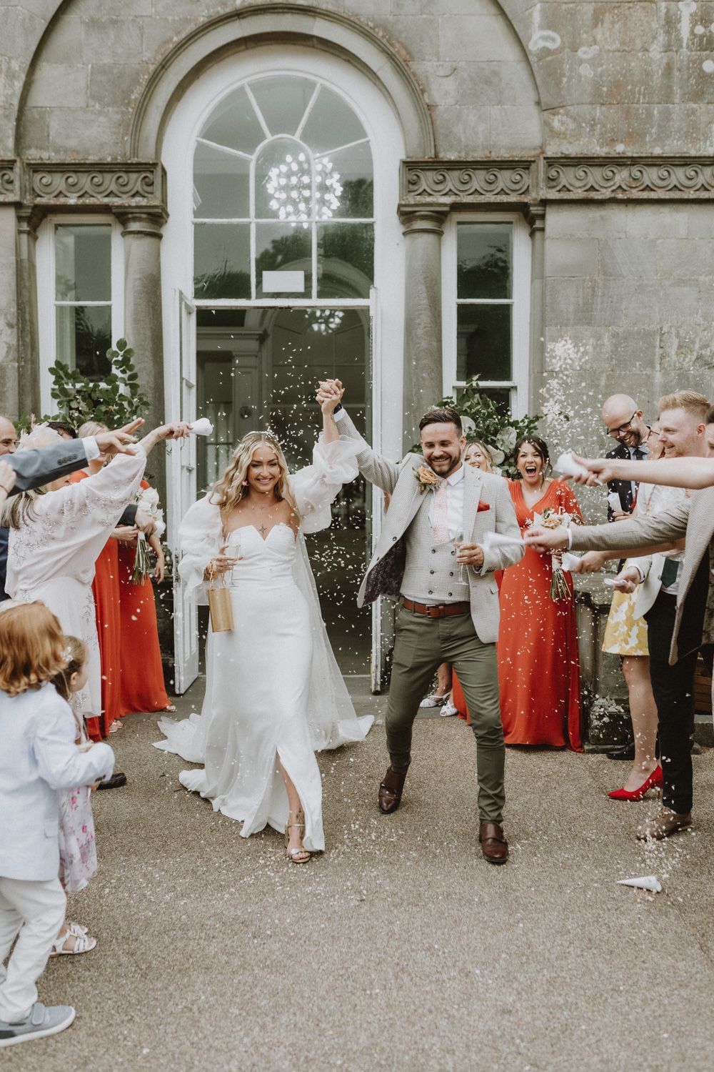 Celebratory moment as wedding guests throw confetti at couple outside stone building with arched windows.
