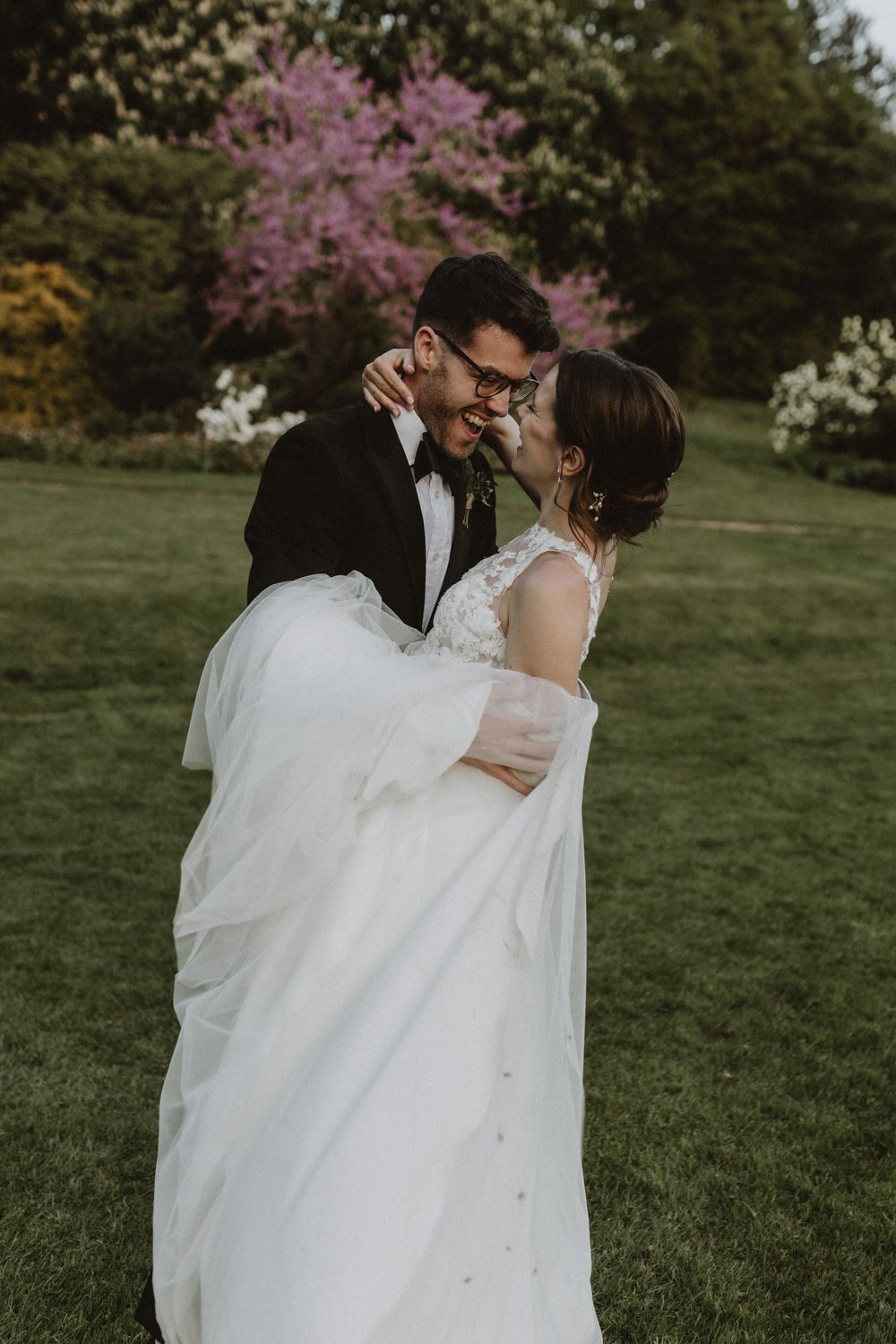 Intimate wedding portrait of couple embracing in garden with blooming cherry blossoms in background.