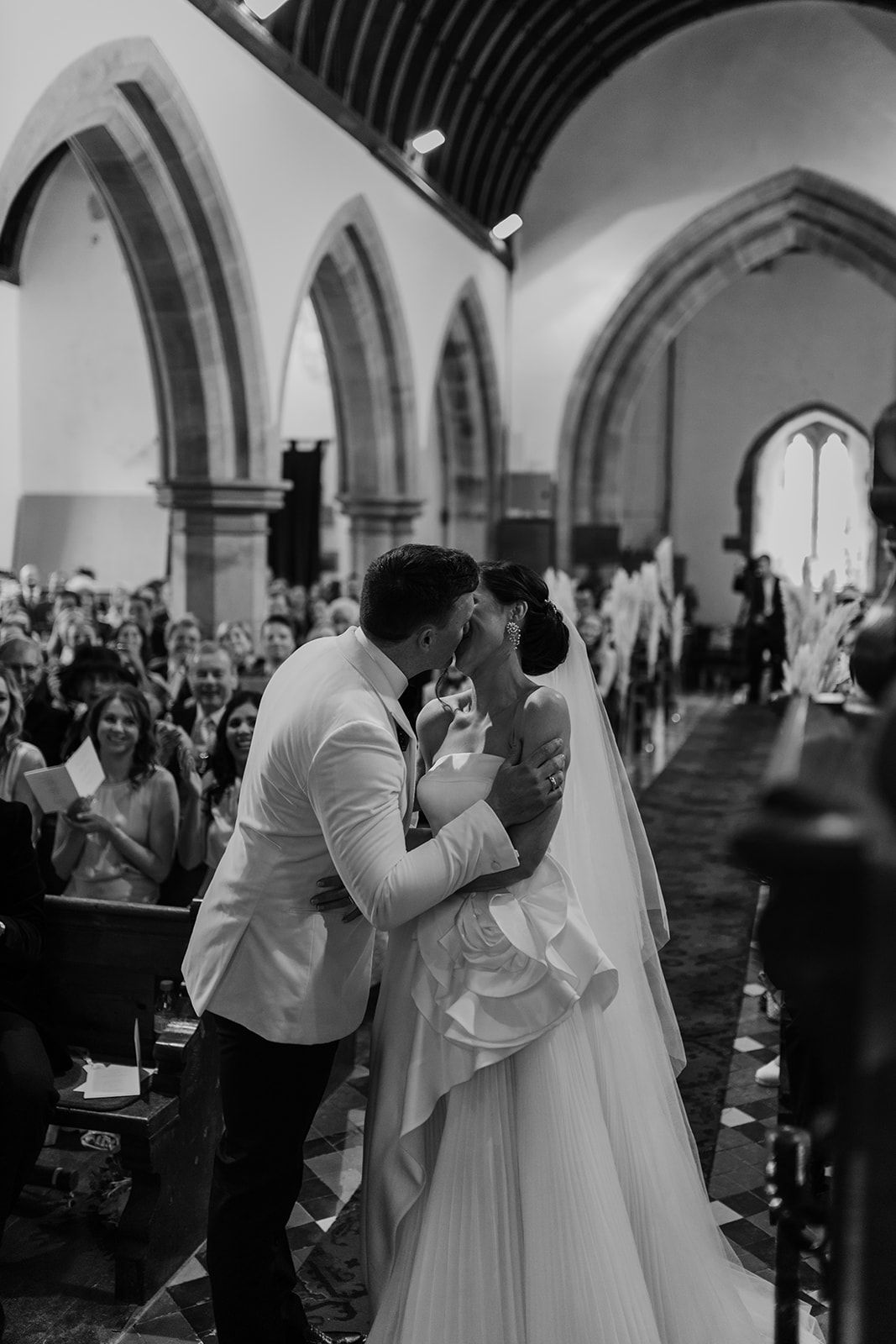 A black and white wedding ceremony photo shows a couple sharing their first kiss in a grand cathedral.