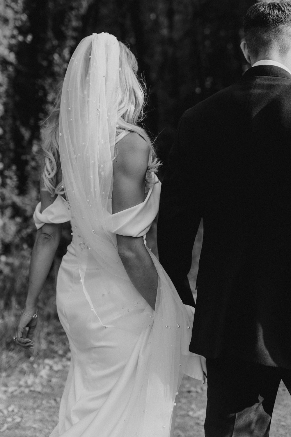Back view of couple in elegant wedding attire walking on a nature path.