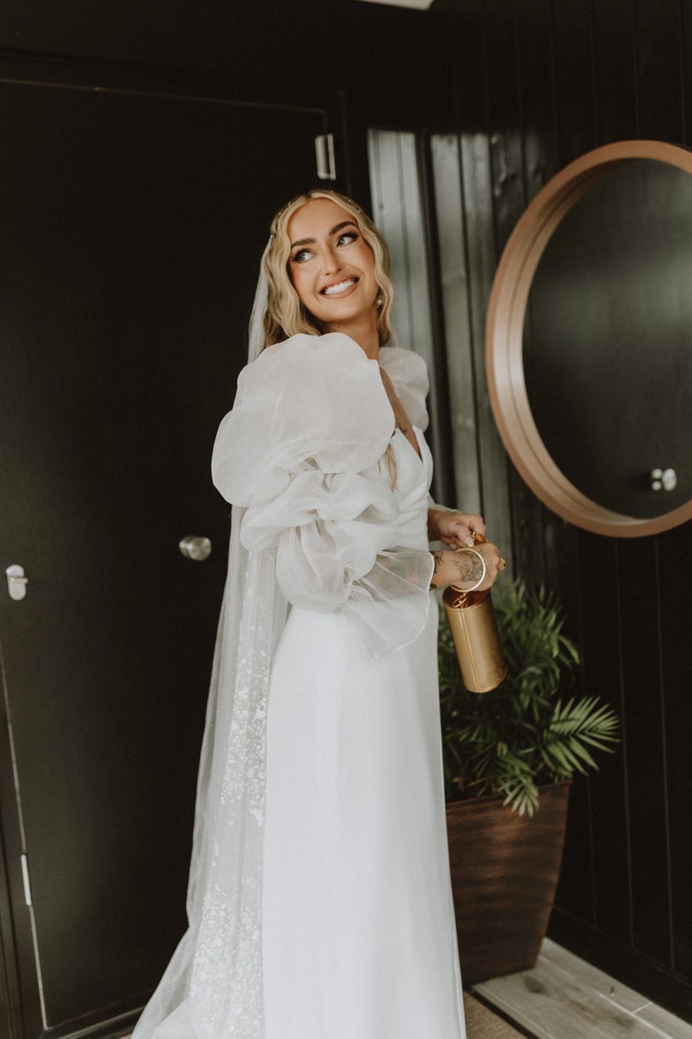 Bride in flowing white dress posing against dark wall with circular mirrors.