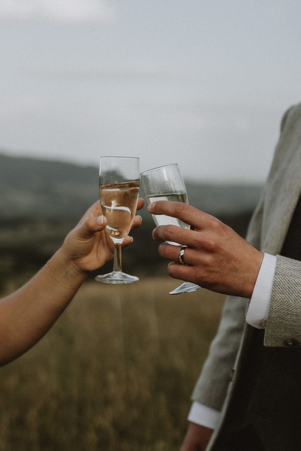 Two hands clink champagne glasses against a scenic countryside backdrop during a wedding celebration.