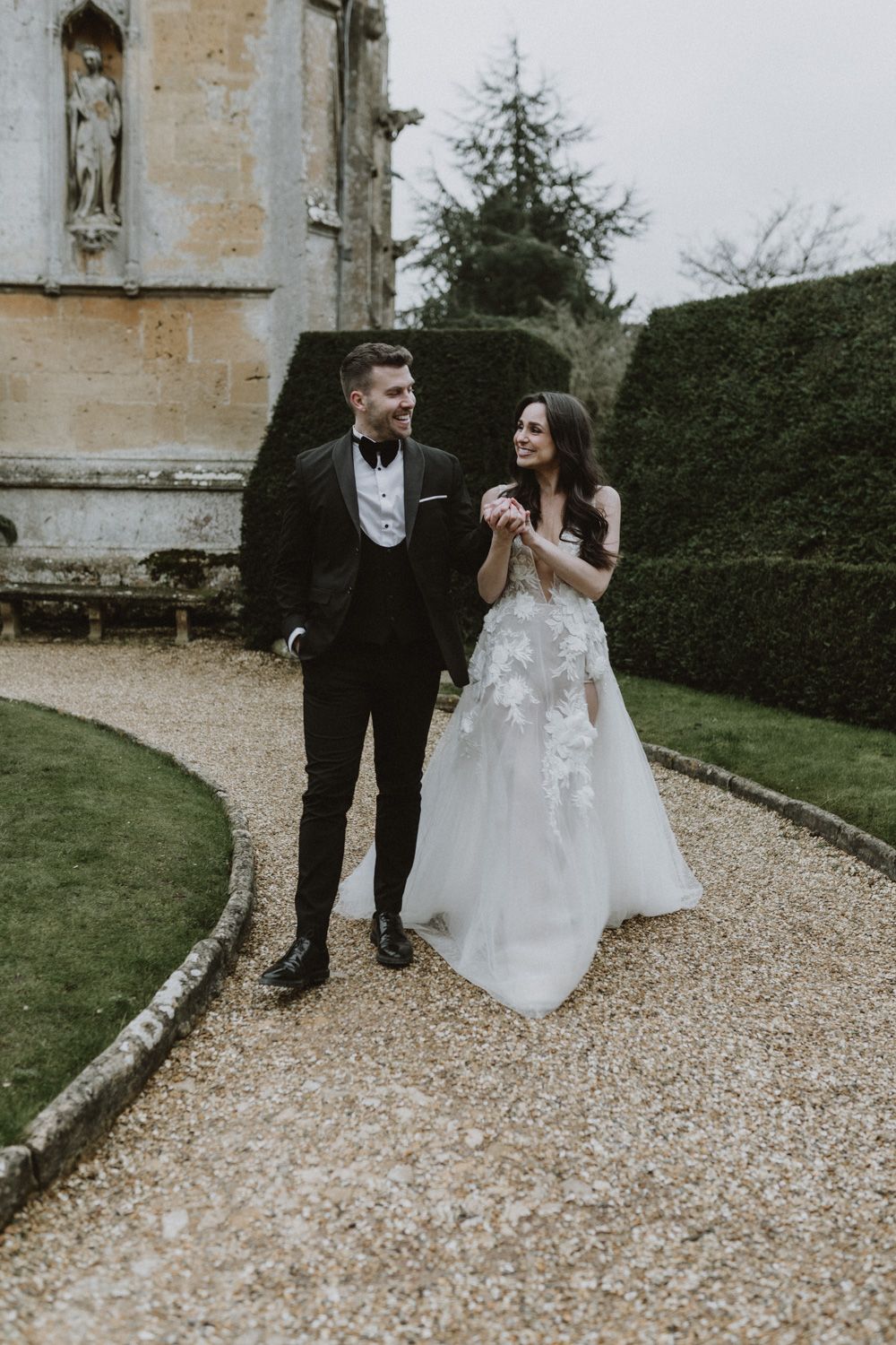 Wedding couple walking along a gravel path outside a historic stone building.