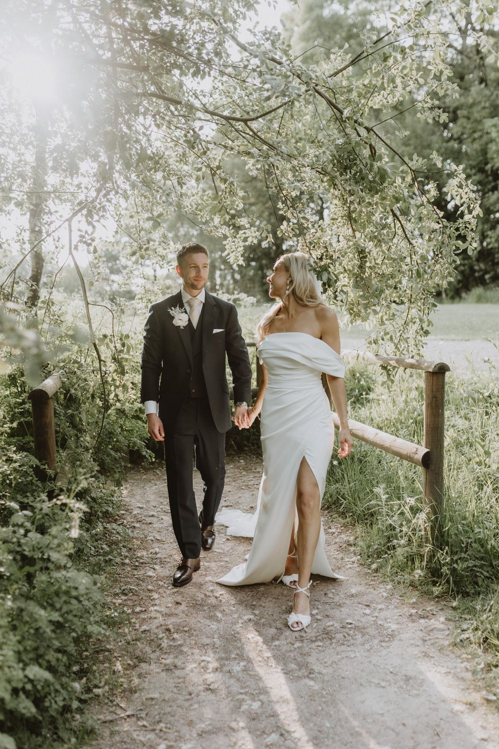 Couple in wedding attire walking along a sunlit garden path under blooming trees.
