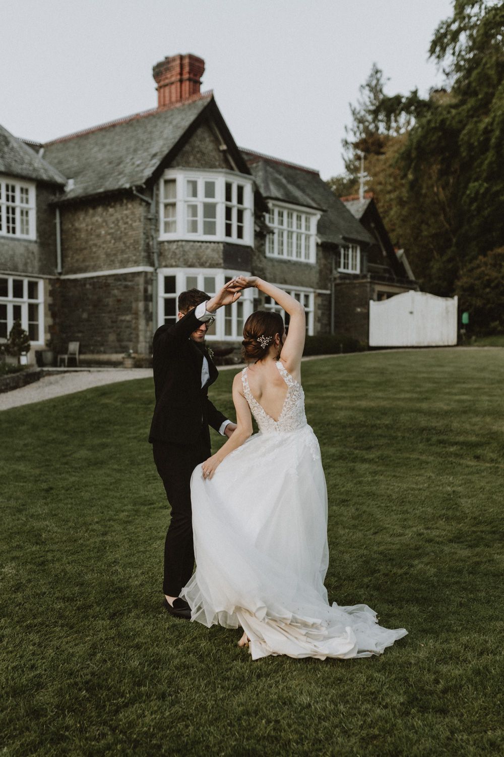Couple dances playfully on a green lawn with a historic stone mansion in the background.